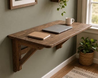 Könnte beinhalten: Ein wandmontierter Schreibtisch aus Holz mit dunkelbraunem Finish, der von zwei abgewinkelten Holzhalterungen getragen wird. Die rechteckige Oberfläche trägt einen geschlossenen silbernen Laptop und ein kleines beiges Notizbuch.
