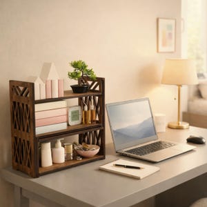 May include: A two-tiered dark brown wooden desktop organiser with intricate lattice-style side panels. The shelves hold a small potted artificial plant, several books with light-coloured spines, a digital clock, glass bottles, and a small bowl.