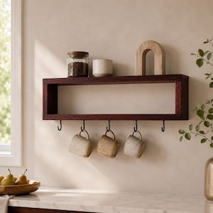 May include: A rectangular dark wood wall shelf featuring five black metal hooks along the bottom edge. Three speckled ceramic mugs hang from the hooks. The top of the shelf holds a glass jar filled with coffee beans, a small ceramic bowl, and a wooden arch.