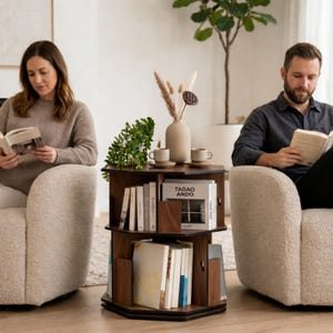 May include: A dark brown, three-tiered, rotating bookshelf with books, a vase, and two small cups. The bookshelf is in a living room setting with two people reading books in cream-colored armchairs. The bookshelf has the text "Tadao Ando" on one of the books.
