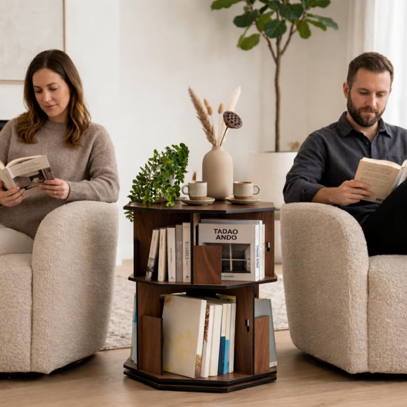 May include: A dark brown, three-tiered, rotating bookshelf with books, a vase, and two small cups. The bookshelf is in a living room setting with two people reading books in cream-colored armchairs. The bookshelf has the text "Tadao Ando" on one of the books.