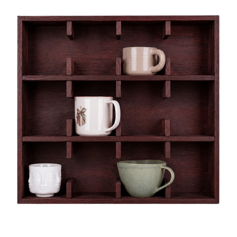 May include: A dark brown wooden wall shelf with a grid of square compartments. Four ceramic mugs are placed inside: a beige mug, a white mug with a small brown graphic, a white mug with a sculpted face design, and a sage green textured mug.