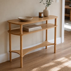 May include: A light wood console table with two oval shelves and four cylindrical legs. The top shelf holds a shallow textured bowl, a small glass candle jar, and a beige vase with greenery. The lower shelf holds two stacked books.