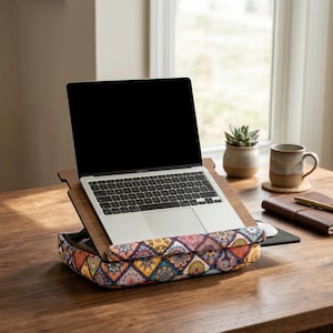 Op de afbeelding: Een laptop staat op een houten schoottafel met een kleurrijk, patroon kussen. Het kussen heeft een geometrisch ontwerp met oranje, geel en blauw. De tafel staat op een houten oppervlak.