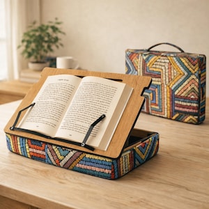 Puede incluir: Un atril de madera con un libro abierto, sostenido por brazos de metal negro. La base del atril y una funda a juego presentan un dise&ntilde;o bordado geom&eacute;trico colorido en tonos azules, amarillos y naranjas. El libro est&aacute; abierto en una p&aacute;gina de texto.