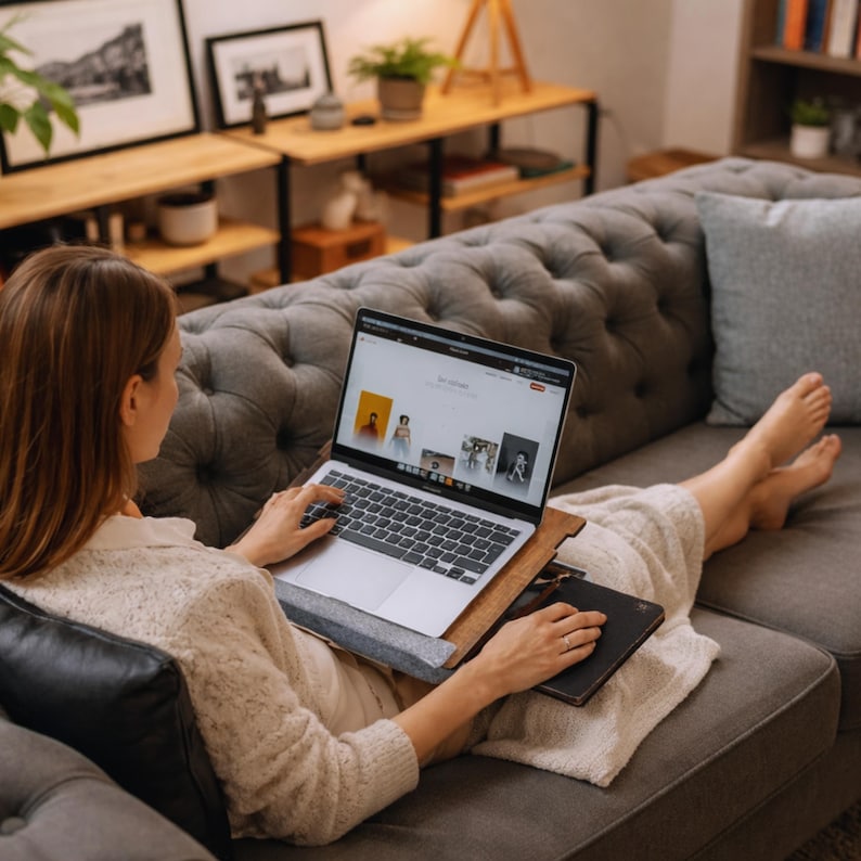 Op de afbeelding: Een persoon gebruikt een laptop op een grijze, gecapitonneerde bank. De laptop staat op een houten en vilten schoottafel, met een zwart notitieboekje ernaast. Het scherm toont een website met verschillende afbeeldingen.