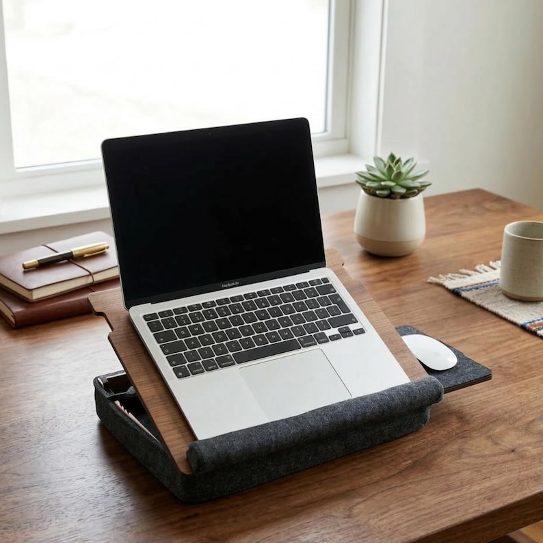 Op de afbeelding: Een laptop op een houten bureau met een grijze vilten laptopstandaard. De standaard heeft een ingebouwde polssteun en een klein compartiment. Een witte muis en muismat bevinden zich rechts van de laptop. Een kleine vetplant in een witte pot is zichtbaar.