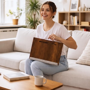 Op de afbeelding: Een houten schoottafel met een handvat en een stoffen kussen. De tafel is warm bruin van kleur en lijkt van hout te zijn. Het stoffen kussen is lichtgrijs van kleur. De tafel is ontworpen om op schoot te worden gebruikt.