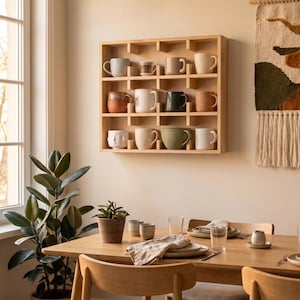 May include: A square light-colored wooden wall shelf with twelve individual compartments, each holding a ceramic mug of varying colors, textures, and shapes. The shelf is mounted on a plain wall above a wooden dining table with place settings.