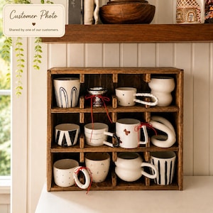 May include: A dark brown wooden cubby shelf containing nine ceramic mugs with various designs, including black stripes, polka dots, and abstract shapes. Some mugs feature unique sculptural handles and are tied with thin red decorative ribbons.