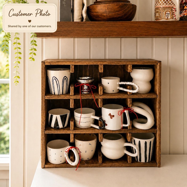 May include: A dark brown wooden cubby shelf containing nine ceramic mugs with various designs, including black stripes, polka dots, and abstract shapes. Some mugs feature unique sculptural handles and are tied with thin red decorative ribbons.