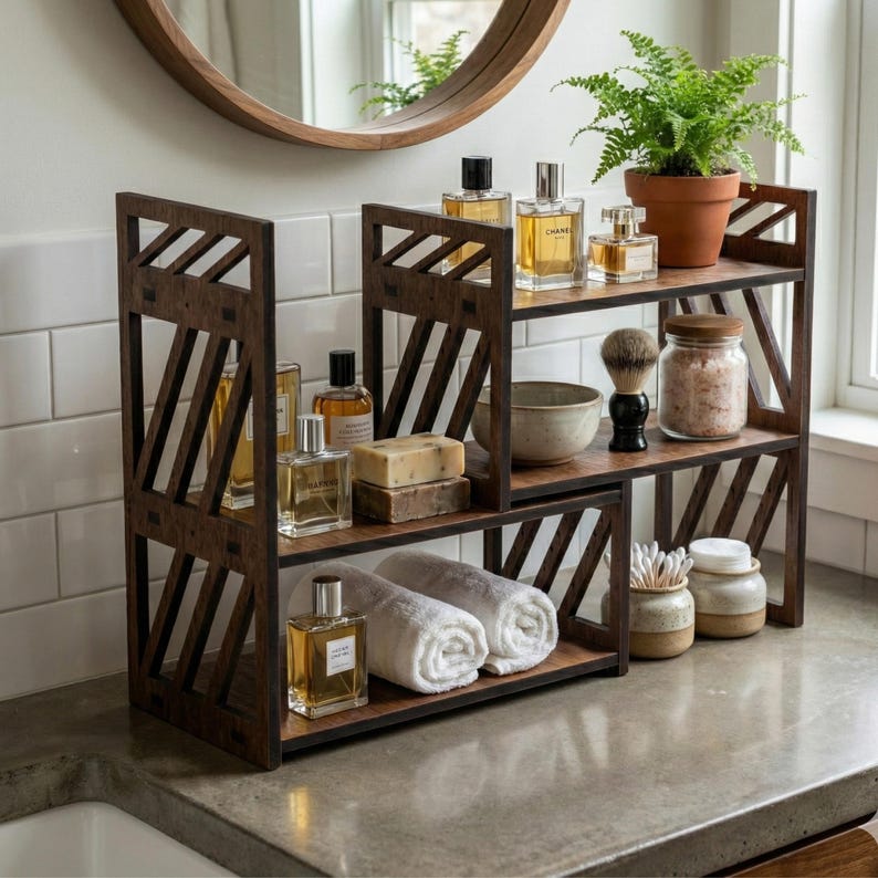 May include: A dark brown wooden tiered bathroom organiser with diagonal side slats. The shelves hold glass perfume bottles, a shaving brush, a ceramic bowl, two white rolled towels, a bar of soap, a small potted fern, and two ceramic jars.