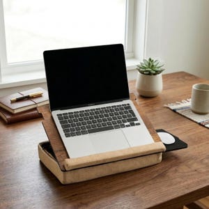 Op de afbeelding: Een laptop staat op een houten laptopstandaard met een ingebouwde muismat. De standaard heeft een beige stoffen polssteun. Een kleine vetplant in een witte pot en een mok staan ook op de houten tafel.