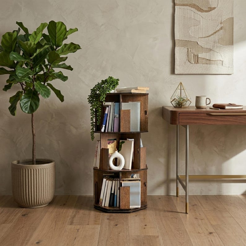 May include: A dark brown, three-tiered rotating bookshelf filled with books and decorative items. The bookshelf is placed on a hardwood floor, with a large potted plant to the left and a desk in the background.