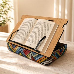 May include: A wooden book stand holding an open book, displaying text from Chapter Seven. The stand features a base with a colourful embroidered geometric pattern in blue, red, yellow, and orange. Black straps secure the book.