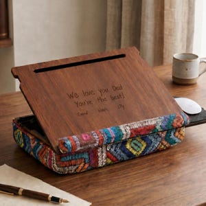 May include: A wooden laptop desk with a hinged top and a colorful embroidered cushion. The top is engraved with the words "We love you Dad, You're the best!" and the names Emma, Noah, and Lily.