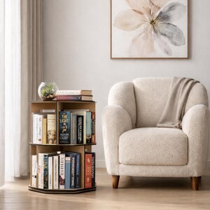 May include: A three-tiered, wooden, rotating bookshelf filled with books. The bookshelf is positioned next to a cream-colored armchair and a floral painting. A small glass terrarium sits atop the bookshelf. The bookshelf is approximately 30 inches tall.