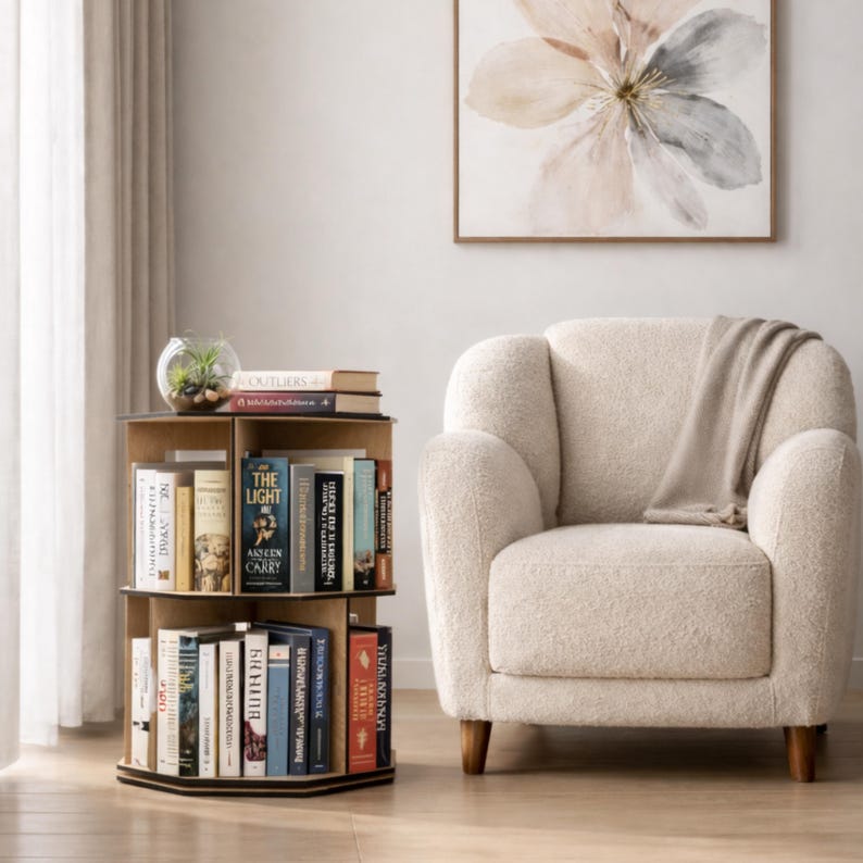 May include: A three-tiered, wooden, rotating bookshelf filled with books. The bookshelf is positioned next to a cream-colored armchair and a floral painting. A small glass terrarium sits atop the bookshelf. The bookshelf is approximately 30 inches tall.