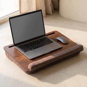 May include: A wooden laptop desk with a brown cushion and a built-in slot for a tablet or phone. A silver laptop and a grey mouse are on the desk. The desk is designed for comfort and convenience.