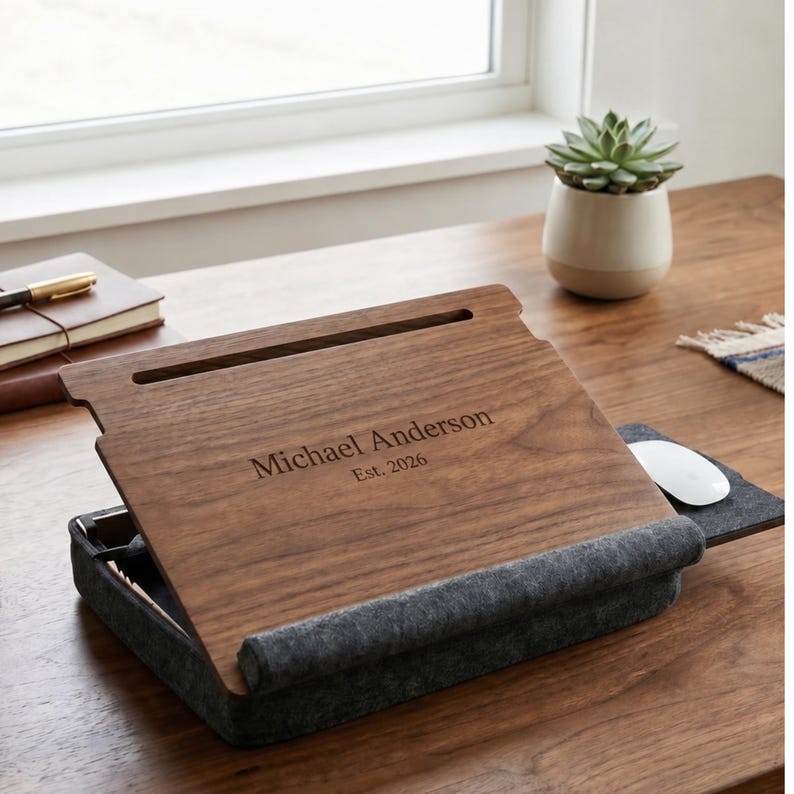 May include: A dark wood laptop desk with a gray felt base and a built-in mouse pad. The wooden surface is engraved with the text Michael Anderson Est. 2026 and features a horizontal slot at the top for holding devices or pens securely in place.
