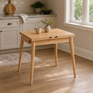 May include: A light-toned wooden dining table with four tapered legs and a rectangular top. The table features a central seam indicating an extendable design, with a small beige ceramic mug and a small potted green plant placed on the surface.
