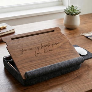 May include: A dark wood laptop desk with a gray felt base and a built-in mouse pad. The wooden surface features a horizontal slot and the engraved text: You are my favorite person. Love, Emma. The desk is angled for use on a flat surface.