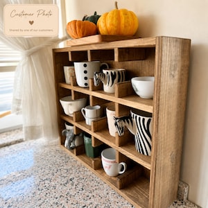 May include: A wooden mug rack with multiple compartments holding various ceramic mugs, including two zebra-patterned mugs with animal head handles. Three decorative pumpkins sit on the top shelf of the brown stained wooden organizer.