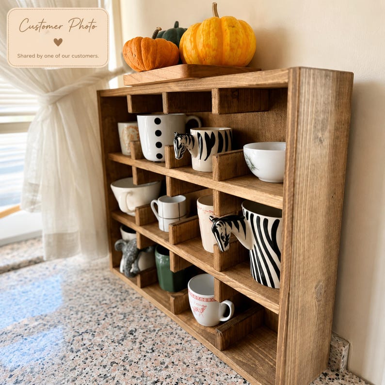 May include: A wooden mug rack with multiple compartments holding various ceramic mugs, including two zebra-patterned mugs with animal head handles. Three decorative pumpkins sit on the top shelf of the brown stained wooden organizer.