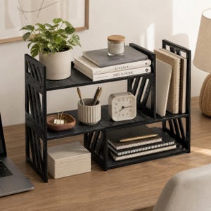 May include: A black wooden desktop organiser featuring two tiers with diagonal side slats. The unit holds a potted plant, books titled KINFOLK and THIS IS HOME, a candle, a square clock, a pen holder, a small wooden bowl, and notebooks.