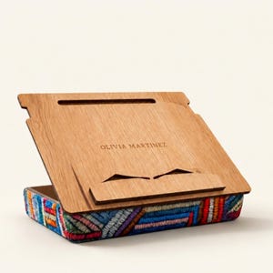May include: A wooden lap desk with a colourful embroidered base. The desk has a hinged top with the name "OLIVIA MARTINEZ" engraved. The base features a vibrant, geometric pattern in various colours.