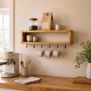 May include: A rectangular light wood wall shelf featuring a top surface for storage and six black metal hooks underneath. Three ceramic mugs hang from the hooks, while a glass jar, a paper bag, and a small bowl sit on the shelf's top surface.
