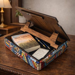 Puede incluir: Un atril de madera con una base bordada colorida. En el interior, un libro titulado "Sweetgrass", un bloc de notas y dos bol&iacute;grafos. El atril est&aacute; abierto, revelando un interior oscuro.