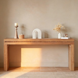 May include: A light brown wooden console table with decorative beading along the top edge. The table is styled with a white ceramic rainbow, a wooden candle holder with a lit candle, a small cork-topped jar, a small white vase with dried flowers, and a tray with decorative items.