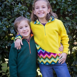 May include: Two children wearing colorful fleece jackets. The girl is wearing a yellow jacket with a rainbow chevron pattern on the bottom. The boy is wearing a green jacket with an orange zipper. Both children are smiling.