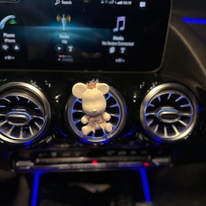 May include: A white and gold bear-shaped car air vent clip. The bear has a crown and bow tie. The clip is placed in the center air vent of a car's dashboard, with a digital display and air vents visible.