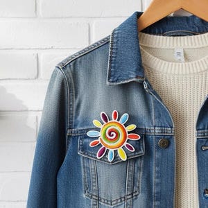 May include: A blue denim jacket with a vibrant, embroidered sun patch. The patch, on the chest pocket, has a spiral centre and petal shapes in red, orange, yellow, green, blue, and purple. The jacket hangs on a wooden hanger.