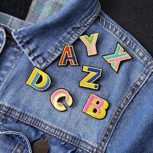 Puede incluir: Primer plano de una chaqueta vaquera azul adornada con parches bordados con letras de colores. Los parches presentan las letras A, B, C, D, Y, Z y un diseño en forma de X, en tonos amarillos, rosas, azules y verdes.