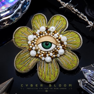 Può includere: Spilla a forma di fiore con un design a occhio centrale. I petali sono verde lime con ricami dorati e l'occhio presenta un'iride verde, circondata da perline bianche, dorate e perlate. Il testo "CYBER BLOOM" è in basso.