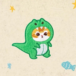 Peut inclure: Écusson brodé représentant un chat mignon déguisé en dinosaure vert. Le chat a un pelage orange et blanc et de grands yeux expressifs. L'écusson est sur un fond beige clair avec des illustrations d'étoiles et de poissons.