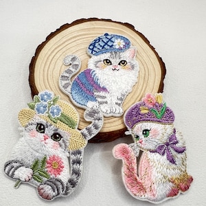 Könnte beinhalten: Drei gestickte Aufnäher mit Cartoon-Katzen. Eine Katze trägt eine blaue Baskenmütze, eine andere einen Strohhut und die dritte einen lila Hut. Jede Katze hat ein einzigartiges Farbschema und ist auf einer Holzscheibe platziert.