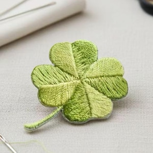 Puede incluir: Un trébol de cuatro hojas bordado en tonos verdes. El trébol es el punto focal, con textura de hilo visible y un ligero brillo. Una aguja e hilo están en primer plano, con tijeras y un pequeño objeto blanco en el fondo.