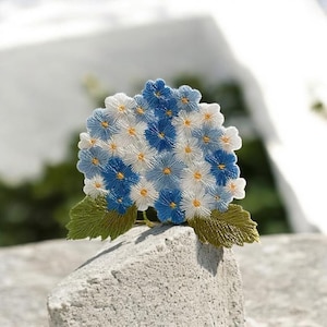 Op de afbeelding: Geborduurde hortensia bloem broche. De broche heeft blauwe en witte bloemen met gele centra en groene bladeren. Het bloemmotief staat tegen een neutrale achtergrond, waarbij de ingewikkelde details van het borduurwerk worden getoond.