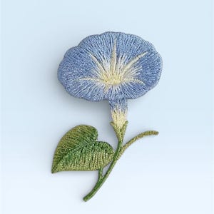 May include: Embroidered morning glory flower with a blue and yellow bloom, green leaf, and stem. The flower has detailed stitching and a textured appearance, set against a light blue background. The design is a floral motif.
