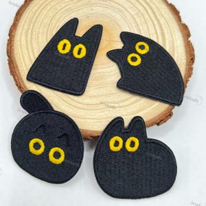 May include: Four black cat-shaped embroidered patches with yellow eyes. The patches are in various poses, including a cat with ears, a cat with a curved body, and a round cat. The patches are on a wooden slice.