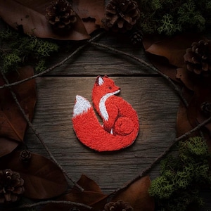 Op de afbeelding: Geborduurde patch van een opgerolde vos met rood haar en een witgepunte staart. De vos is geplaatst tegen een rustieke houten achtergrond, omringd door bladeren, dennenappels en mos. Een gedetailleerd, handgemaakt item.