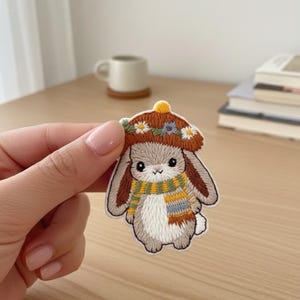 May include: Embroidered patch of a bunny wearing a brown hat with floral details and a striped scarf. The bunny has brown ears and is in a seated position. The patch has a white background and a stitched border.
