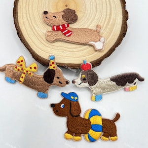 Könnte beinhalten: Gestickte Dackel-Patches in verschiedenen Designs. Ein Hund trägt einen roten Schal, ein anderer eine Partyhütchen und ein dritter eine blaue Mütze und einen Schal. Die Patches sind braun, hellbraun und weiß mit blauen und gelben Akzenten. Die Patches befinden sich auf einer Holzscheibe.