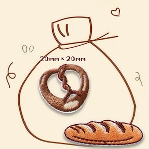 May include: Embroidered patches of a pretzel (2cm x 2cm) and a loaf of bread (3cm x 1cm). The patches are brown and tan, with detailed stitching. The design includes a drawing of a bag and decorative elements.