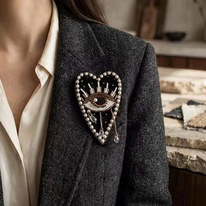 Puede incluir: Broche en forma de corazón con diseño de ojo. El broche es negro, con cuentas blancas y doradas alrededor del ojo y el borde del corazón. Dos cristales transparentes en forma de lágrima cuelgan de la parte inferior. El broche está prendido a una blazer gris oscuro.