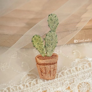 May include: Embroidered cactus in a brown pot. The cactus is green with detailed stitching, and the pot is a warm brown. The design is set against a backdrop of delicate white lace and sheer fabric, creating a soft, textured look.